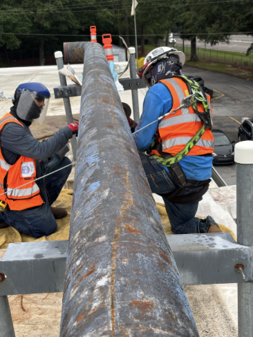 Two workers in high-visibility vests and hard hats installing a large rusty pipe on a commercial rooftop