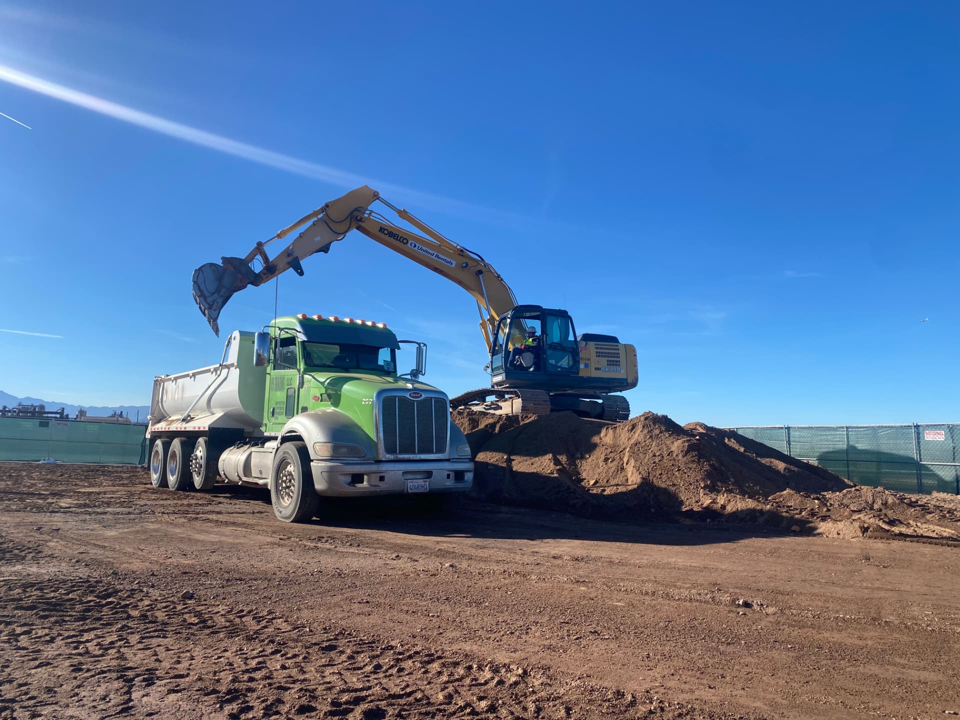 Kobelco excavator loading a dump truck during mass excavation on a fenced commercial site