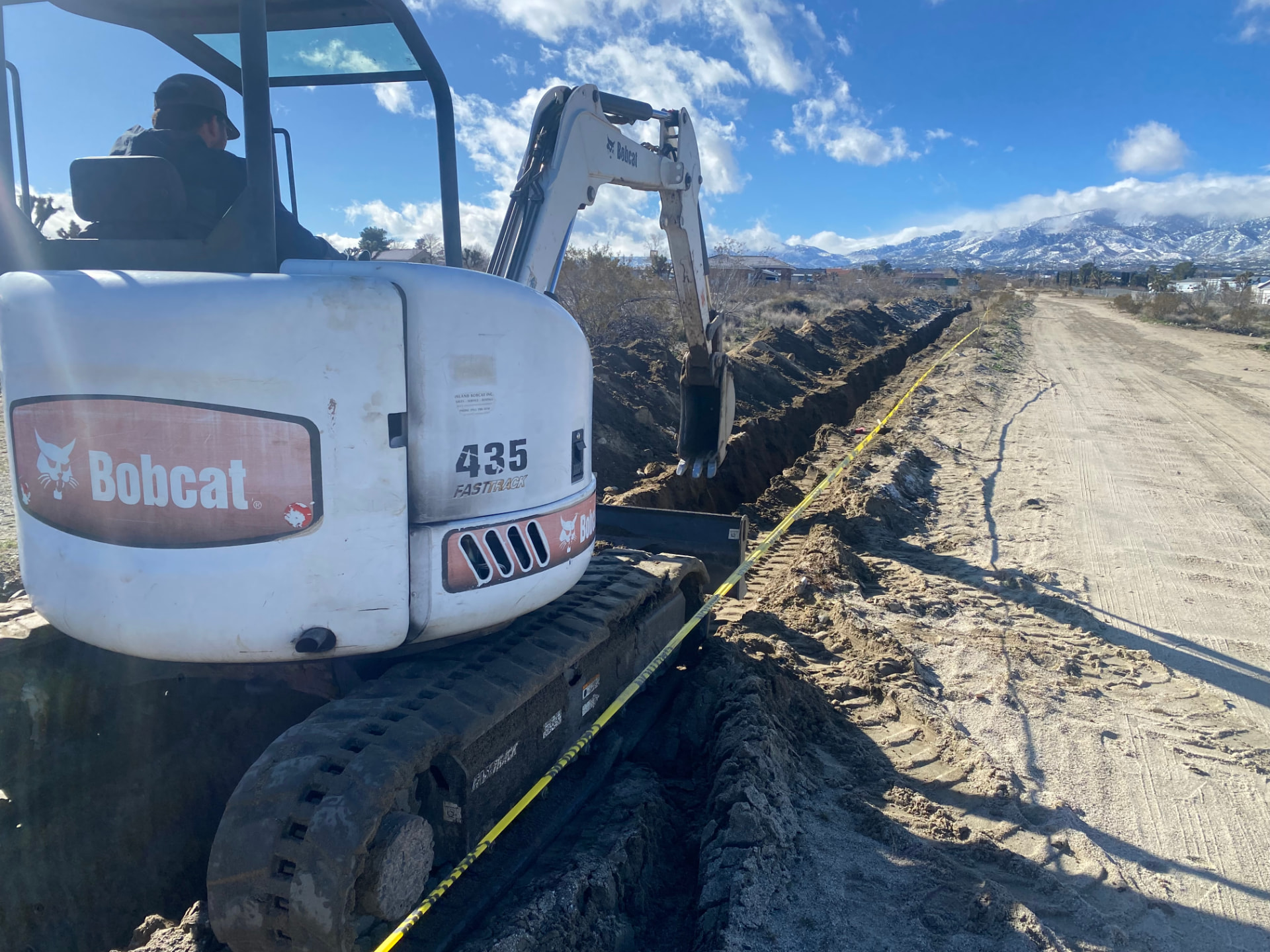 Bobcat 435 excavator cutting a utility trench on a desert jobsite with snow-capped mountains behind